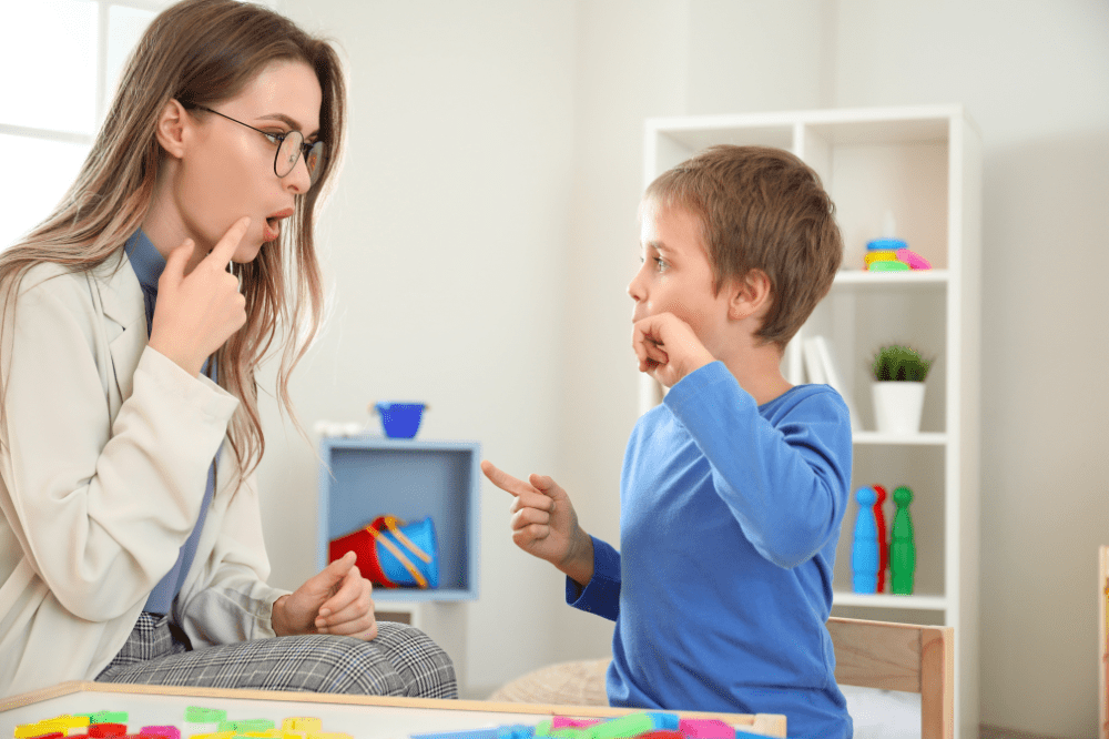 A female speech therapist demonstrates mouth movement by touching her chin as a young boy mimics the gesture at a table with colorful toys.