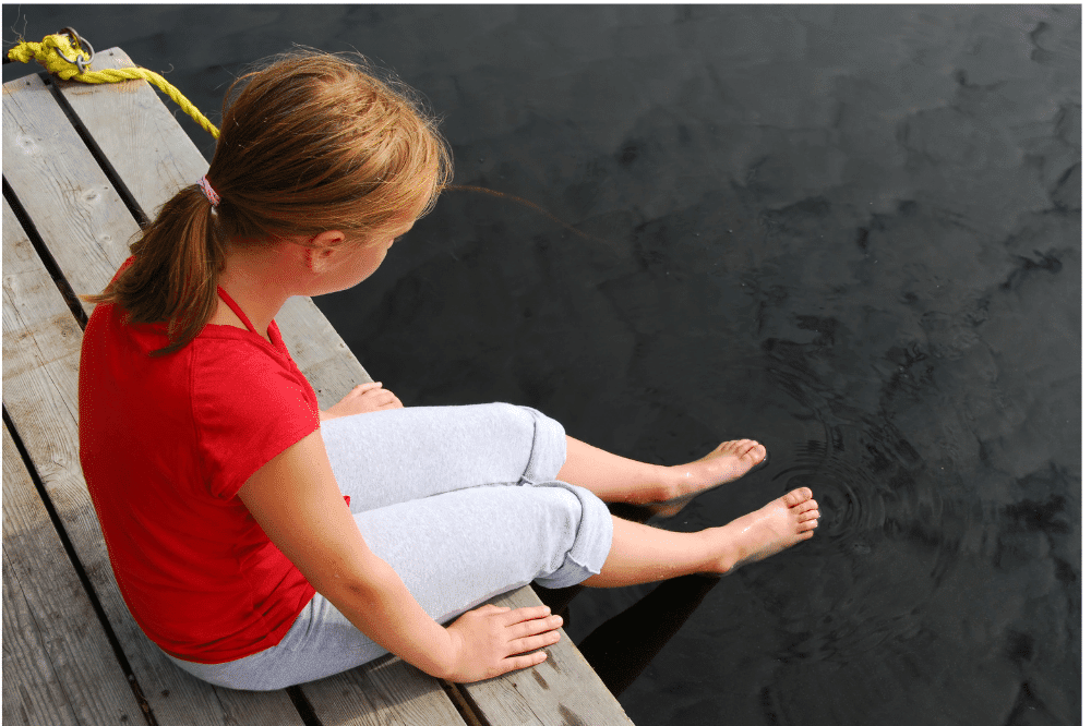 A young girl on a dock looks thoughtfully at the water, contemplating her first day of school and the journey to come.