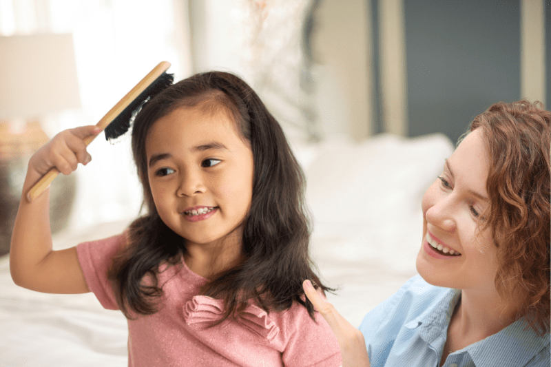 Child brushing their own hair with parental supervision to build self-care and school readiness skills.